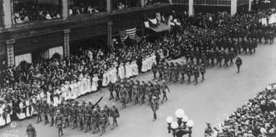 American troops march in a parade down a city street with many onlookers on the sidewalks.