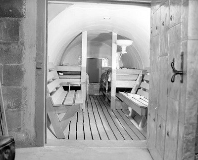 looking through a door into an underground bomb shelter with vaulted ceiling, wooden floor, two wooden benches facing each other, and two wooden beds side by side. One light on a post illuminates the room.