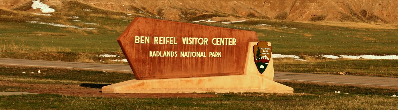 a sign reading ben reifel visitor center sits in front of a road and badlands formations, all bathed in a warm sunset glow.
