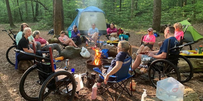 A group of people, some in GRIT freedom wheelchairs sitting around a campfire in a circle.