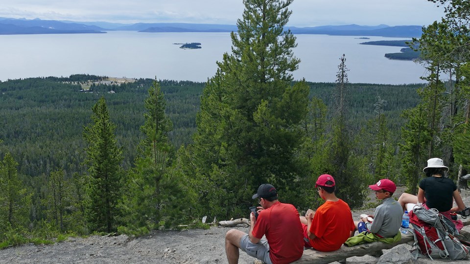 Four hikers sit on a rock at the top of the mountain, with a view of the lake in the distance.