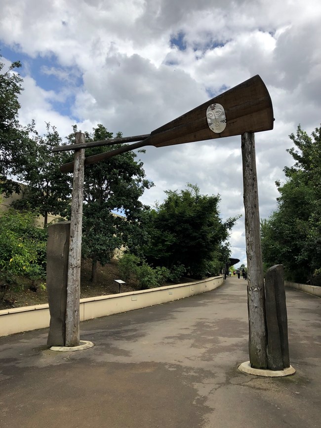 A gate with a glass mask and crossed canoes leads to a walking path.