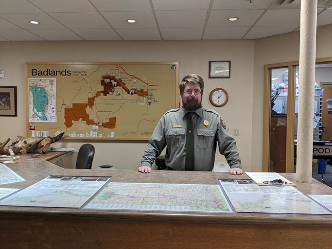 a bearded park ranger smiles from behind a tall desk with a map of the park on the wall behind him.