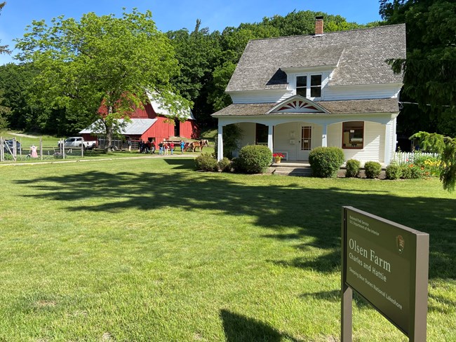 A white farmhouse in the foreground with people standing next to a barn and horses in the background.