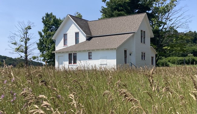 Amongst seeding green grass, a white farmhouse with red trimmed windows, a bright blue sky and several trees poking out from behind the house.