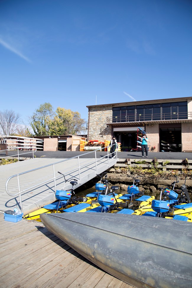 A two story building with an angled deck that leads to a waterfront area where canoes and other boats are stored.