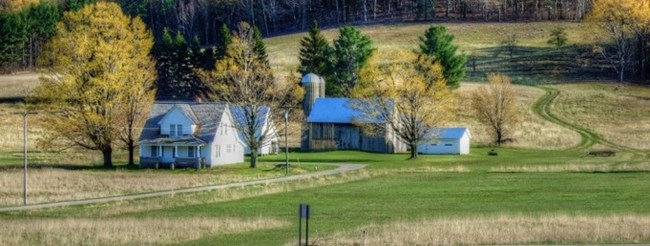 Dechow's (Deco) 2-story house with a barn behind with towering silos surrounded in changing almost leafless trees and long yellowing grass.