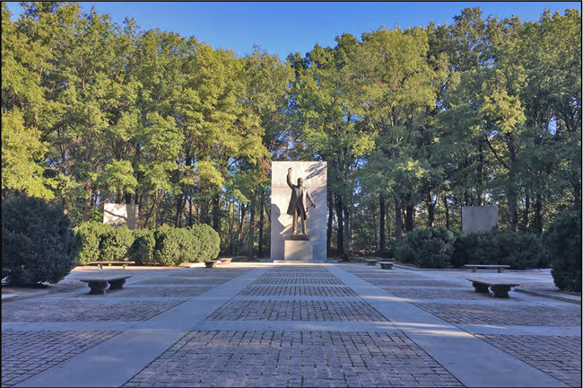 In the background and in the distance stands the statue of Theodore Roosevelt dressed in a suit raising his right hand in the air standing in front of a taller concrete backing.