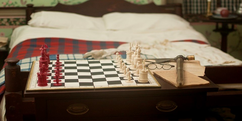Color photo showing a replica of an 18th-century chess set and glasses on a wooden dresser at the foot of a wooden bed.