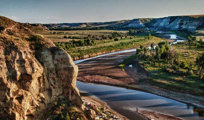 Winding river with red rock embankments and valley of green trees with buttes on either side.