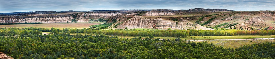 The Little Missouri River stretches across a grassy and wooded river bottom with bare, striped buttes rising up beyond the far bank.
