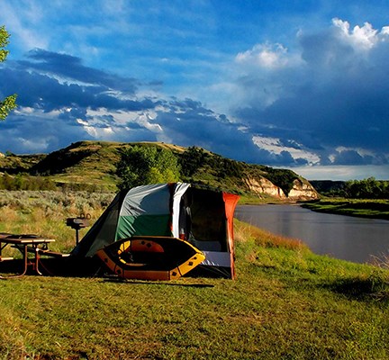 A brightly colored tent sits on a bank above the Little Missouri river with sun-bathed buttes in the background.