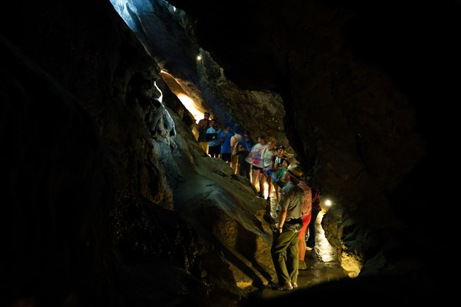 A narrow ambient lit cave passage with multiple visitors and a ranger