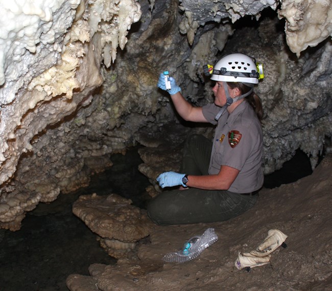 Woman wearing NPS uniform, a hard hat, and latex gloves sits next to a cave pool, holding a sample bottle of water up to the light.