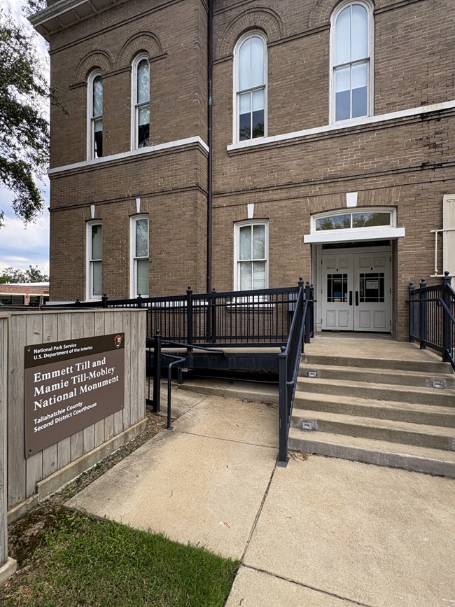 Two glass doors lead into a brick building with a cement ramp and several stairs. A sign reads "Emmett Till and Mamie Till-Mobley National Monument"