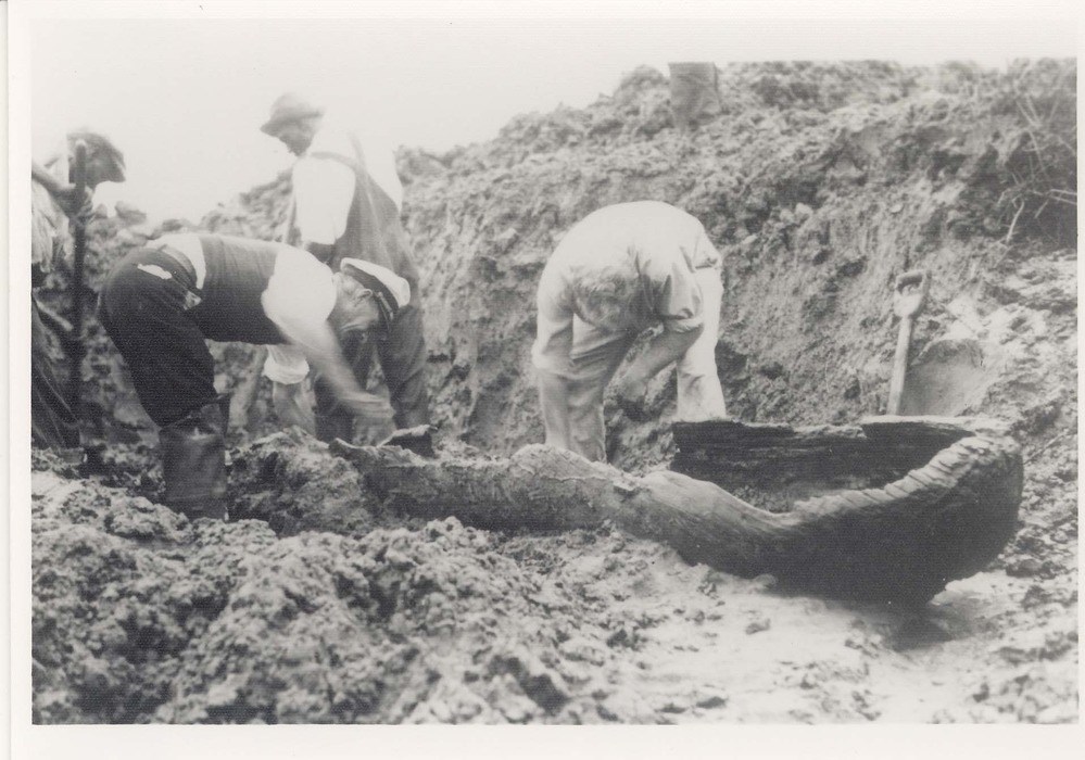 black and white historic photo of people digging up a wooden canoe