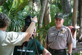 Image of person taking photo of park ranger. One person ducks below camera and one person seen in background. Background filled with palm trees and vegetation.