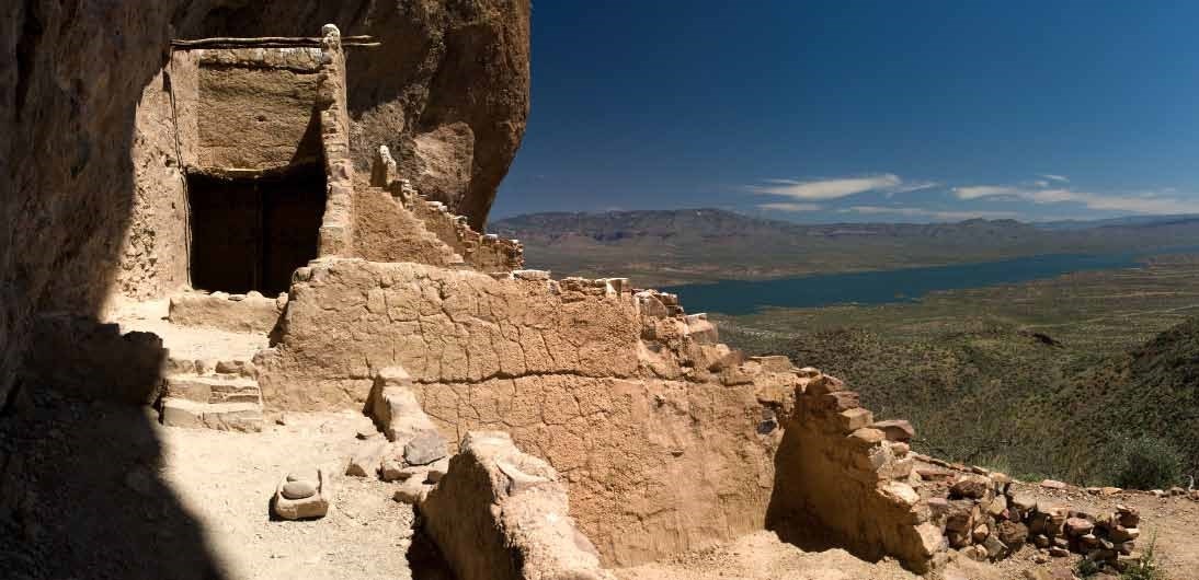 Image of multiple cliff dwelling rooms with Roosevelt Lake and mountains in the background.