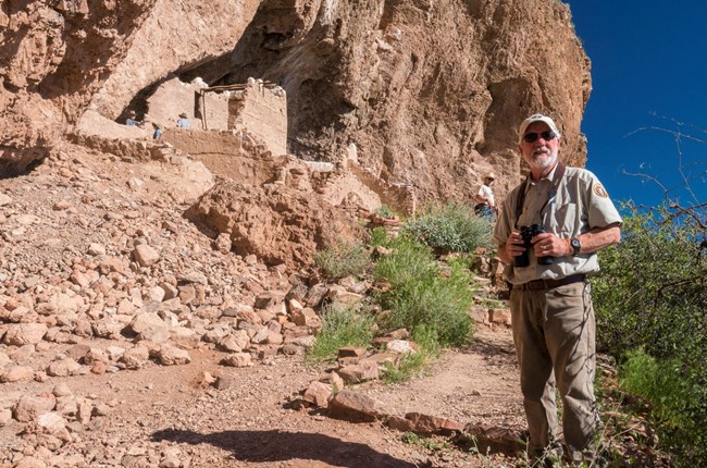 Park volunteer with binoculars looks out. Upper Cliff Dwelling seen in the background.