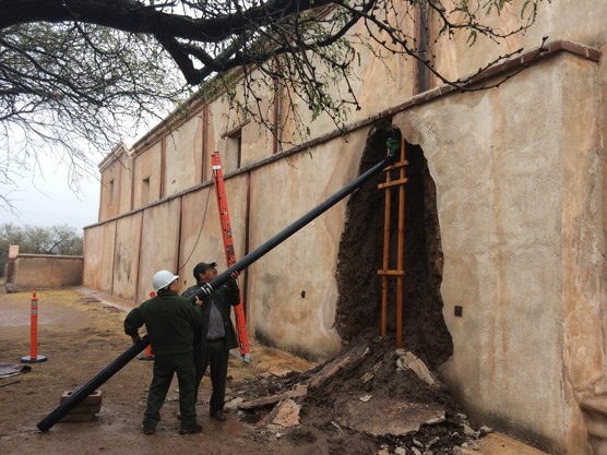 two men hold up a large pvc pipe to catch rain runoff from adobe wall collapse