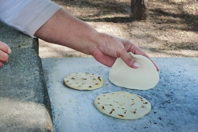 Corn tortilla being place on a hot comal with two others ready to be taken off.