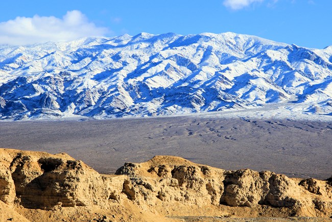 A snow-covered mountain range stands tall behind tan badlands in a desert landscape.