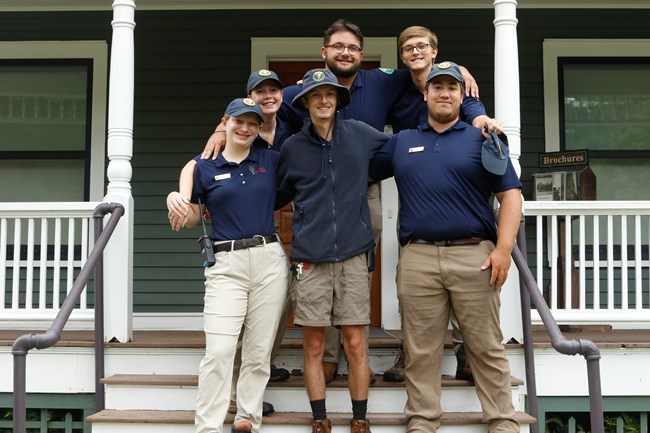 Group of six young men and woman on the steps of a house in navy volunteer uniforms. They stand with their arms around each other.