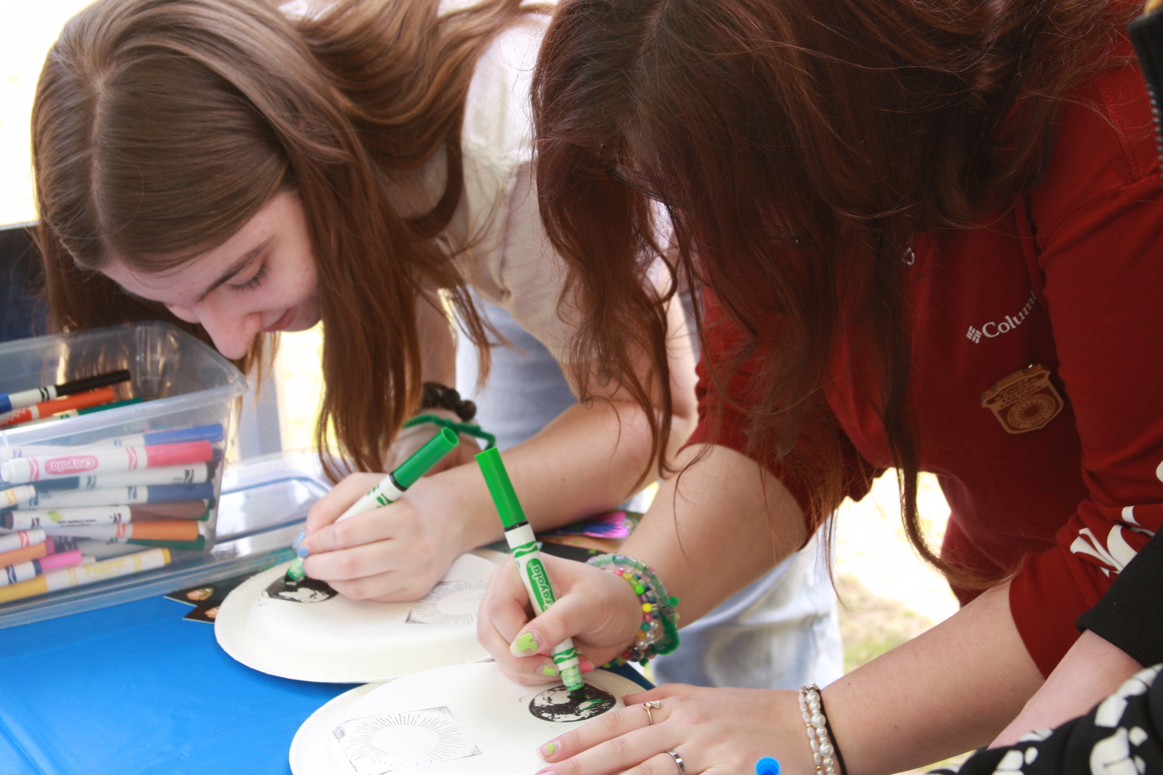 Two teenage girls hunch over table, using markers to color and draw on paper plates.