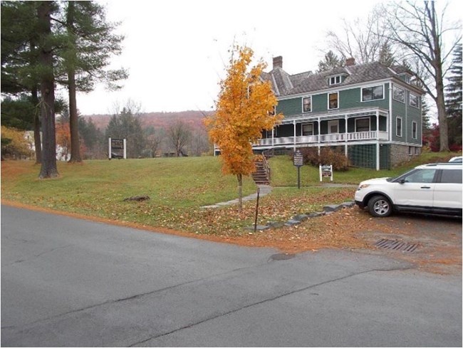 view of Zane Grey Museum from the paved road in front. Museum is large 2-story green building on slight grassy hill. Steps lead up to the house. White cars are parking on the right.