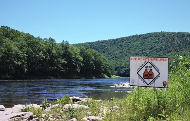 view of river. Sign next to river reads "Life Jackets Save Lives" with a graphic of a life jacket and NPS arrowhead. More text reads "Swimming, boating, fishing, floating. Wear It!"