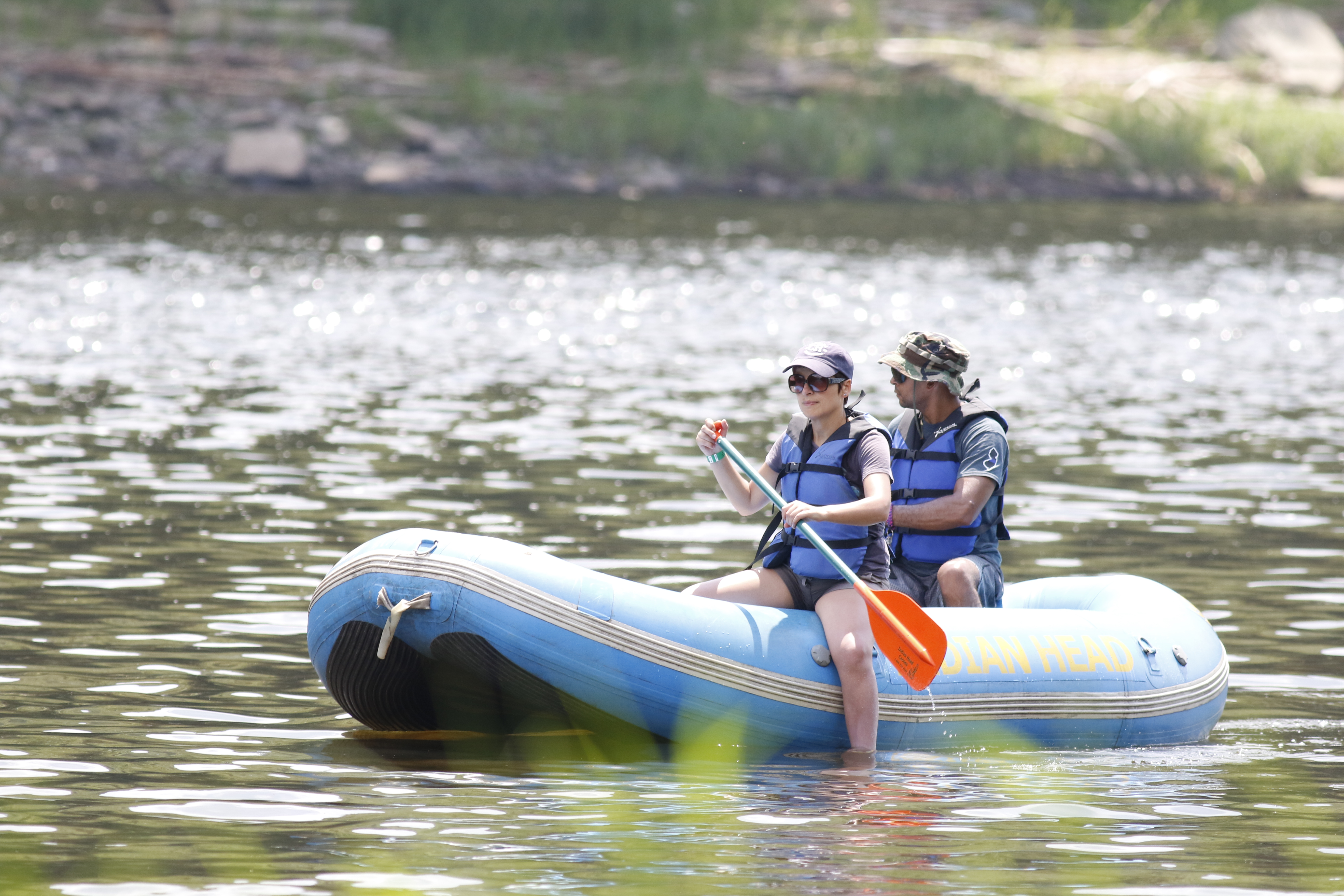 two people in hats, sunglasses, and lifejackets sit atop a blue inflatable raft in water. Person sitting in front holds a blue and orange padde.