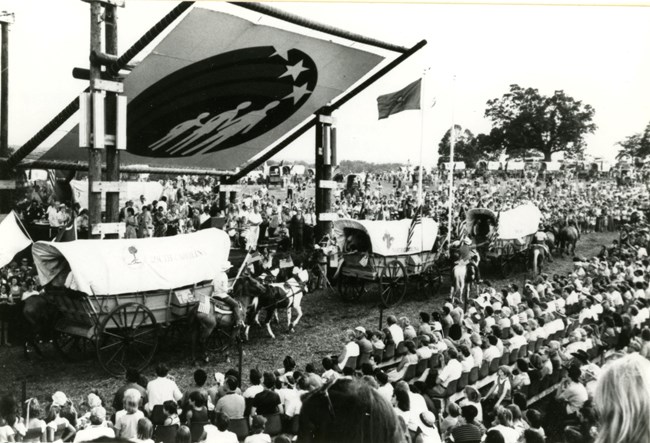 black and white photo of a large crowd seated in chairs in front of a stage and a line of conestoga wagons