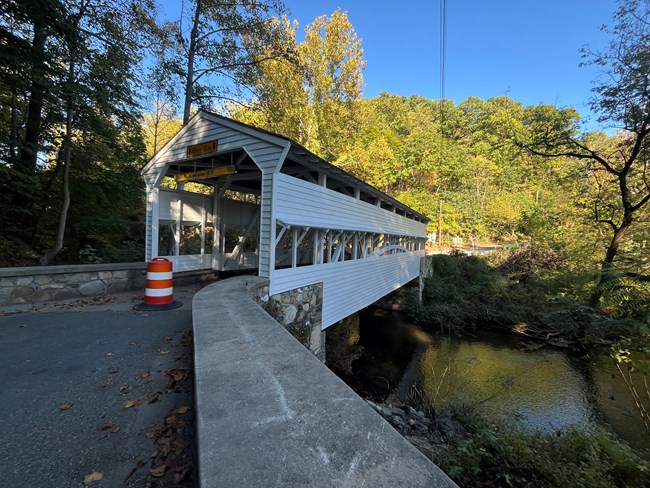 A covered bridge with wooden siding painted white spans a creek.