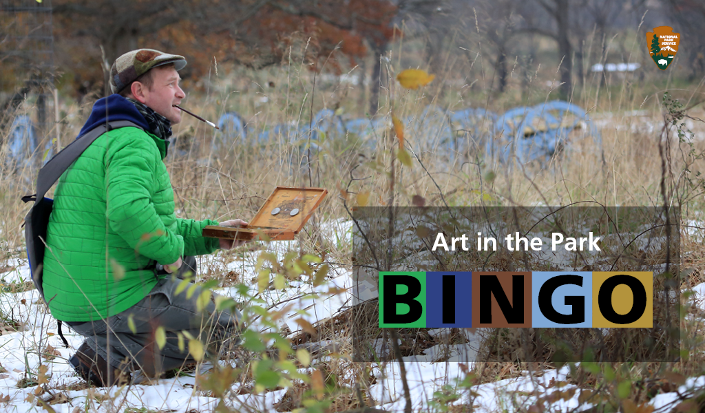 a man with paintbrush in his mouth crouches in a field with cannons in the background, text reads Art in the Park Bingo