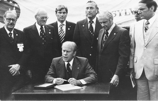 A black and white photo of President Gerald Ford seated at a small wooden table surrounded by men in suits