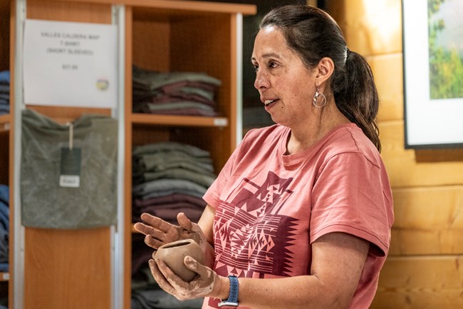 A woman holds up a small, handmade pot while speaking to a crowd.