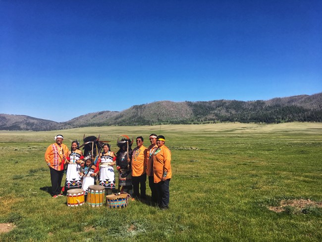 A group of Pueblo singers and dancers dressed in traditional regalia stand in a montane grassland.
