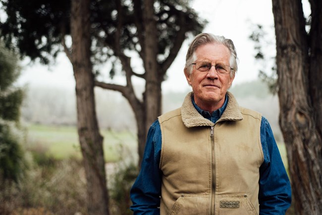 A gray-haired man wearing glasses stands in a forest.