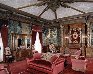 A bedroom with tapestry wall coverings and richly carved ceiling.