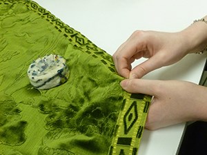 A pair of hands sewing silk braid onto a patterned velvet.