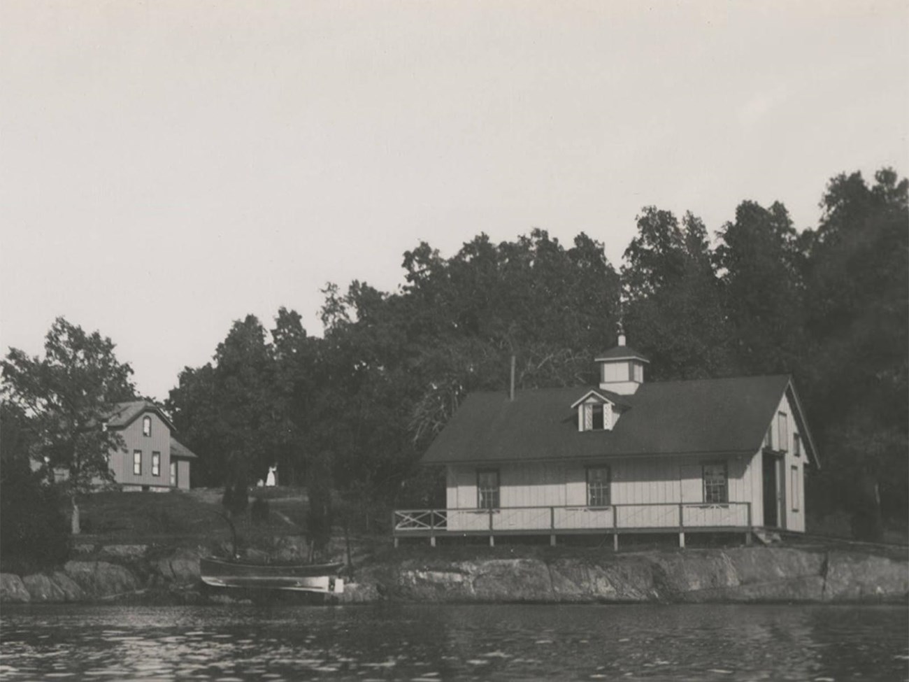 A black and white photograph of a wooden building on the shore of a river.