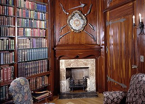 A room with bookshelves lining the walls and a stone fireplace with antique firearms hung above.
