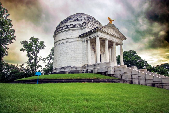 Large memorial stone structure with a domed roof and front pediment. Row of stone steps leading up to entrance. Dark clouds in sky above memorial.