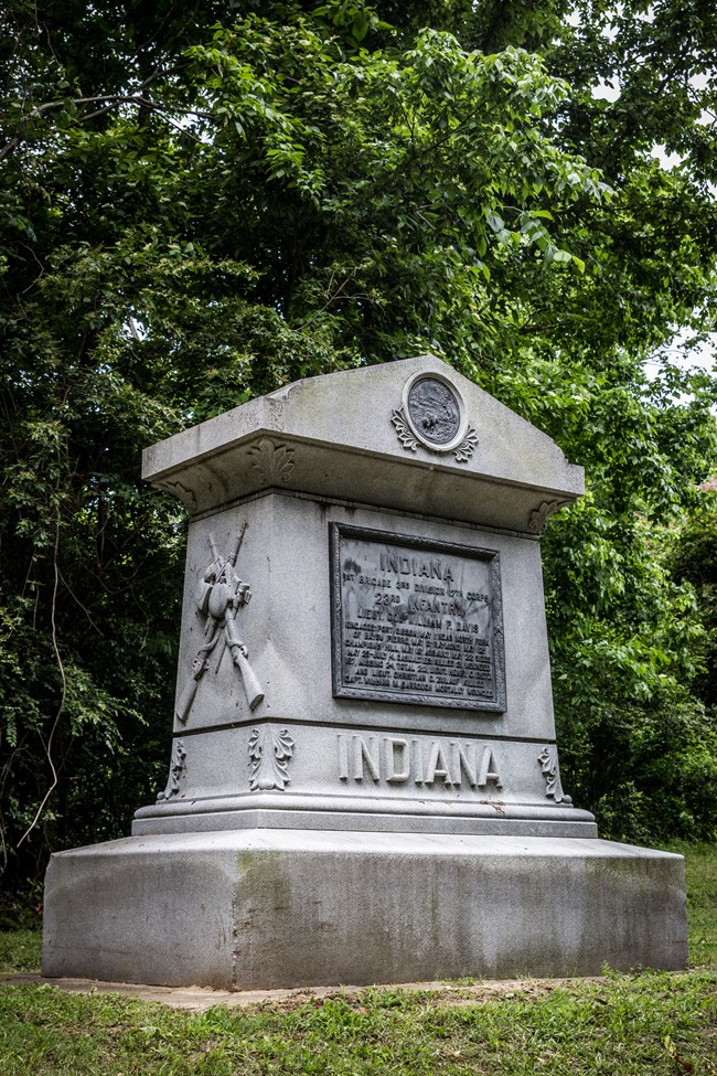 A granite marker with a bronze plaque with an inscription written on the front. On the side of the monument carved into the stone are two weapons