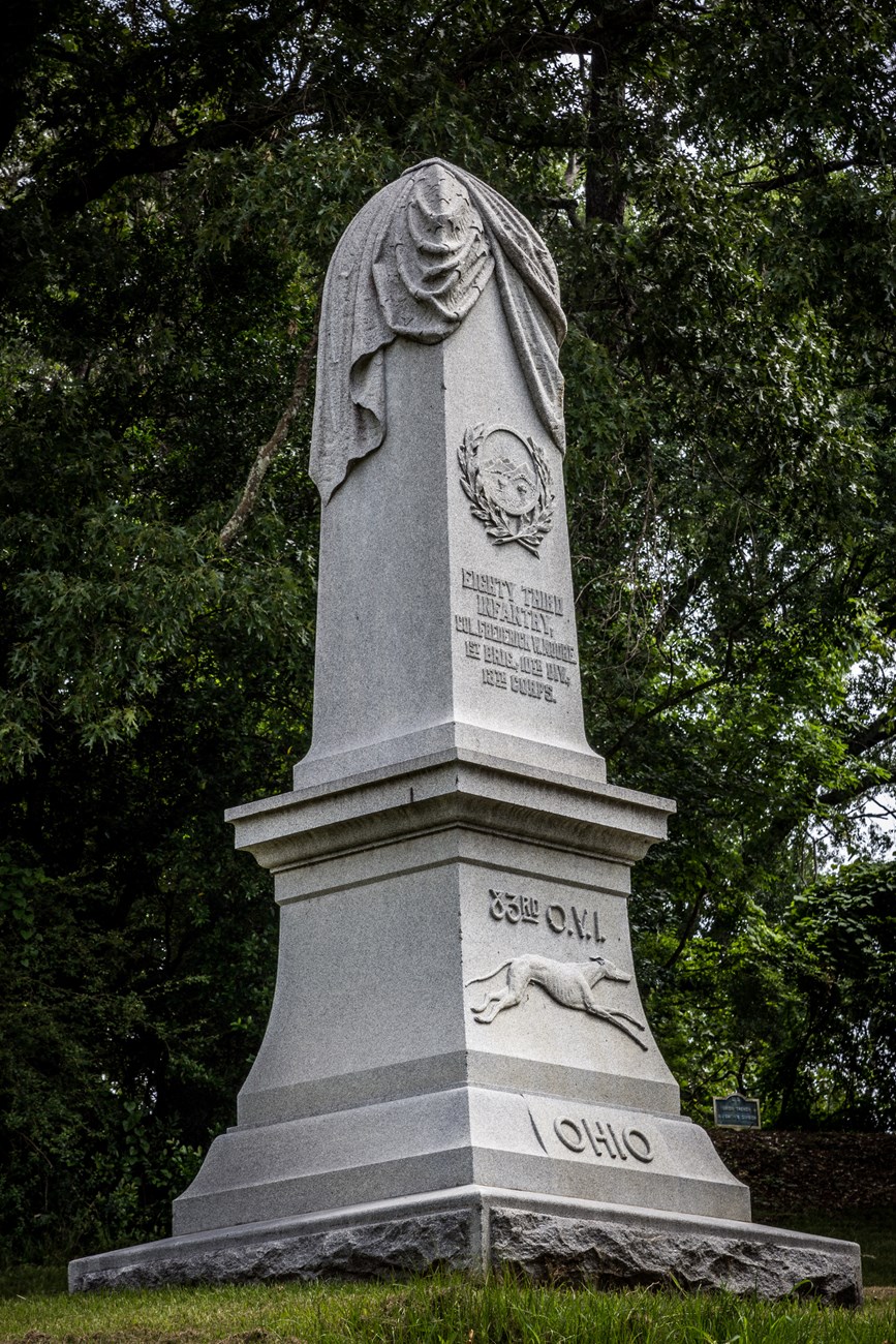 A tall vertical statue sits on a hill with a short inscription on the front of it