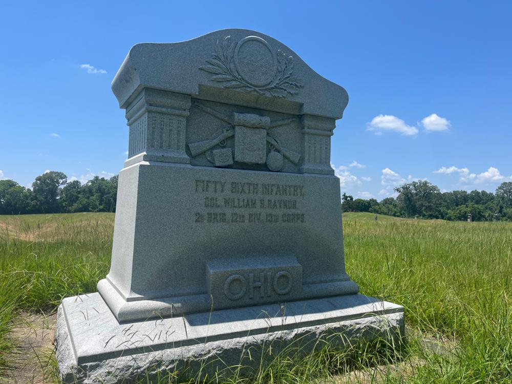 Square stone monument with rounded top, framing a display of 2 crossed rifled muskets behind a knapsack. Inscription: Fifty-Sixth Infantry. / Col. William H. Raynor. / 2d Brig., 12th Div., 13th Corps. / OHIO