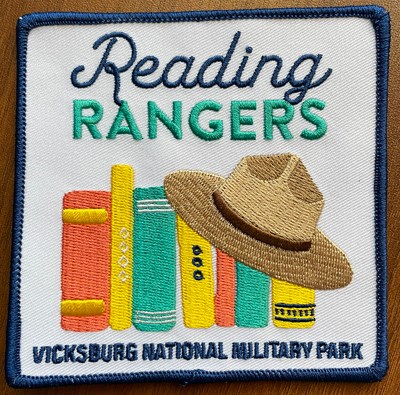 Patch with royal blue embroidered border and white background. Reading Ranger written across the top above a row of colorful books with a park ranger hat covering the right corner of the row. Vicksburg National Military Park written below the books