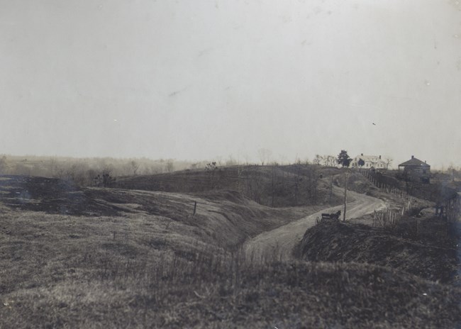 A black and white image showing a rolling landscape without trees, two houses on rise, and a road.