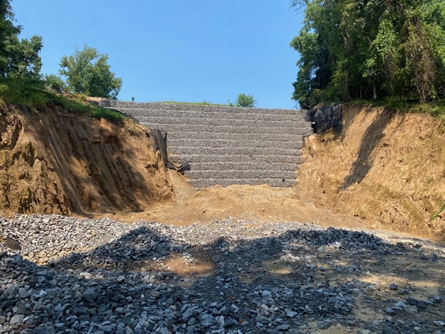 Center of image is very tall wall made up of gray stone in wire rectangles. On either side of the wall are light brown exposed earthen cliff faces. Blue sky above with trees grown on top of cliffs.