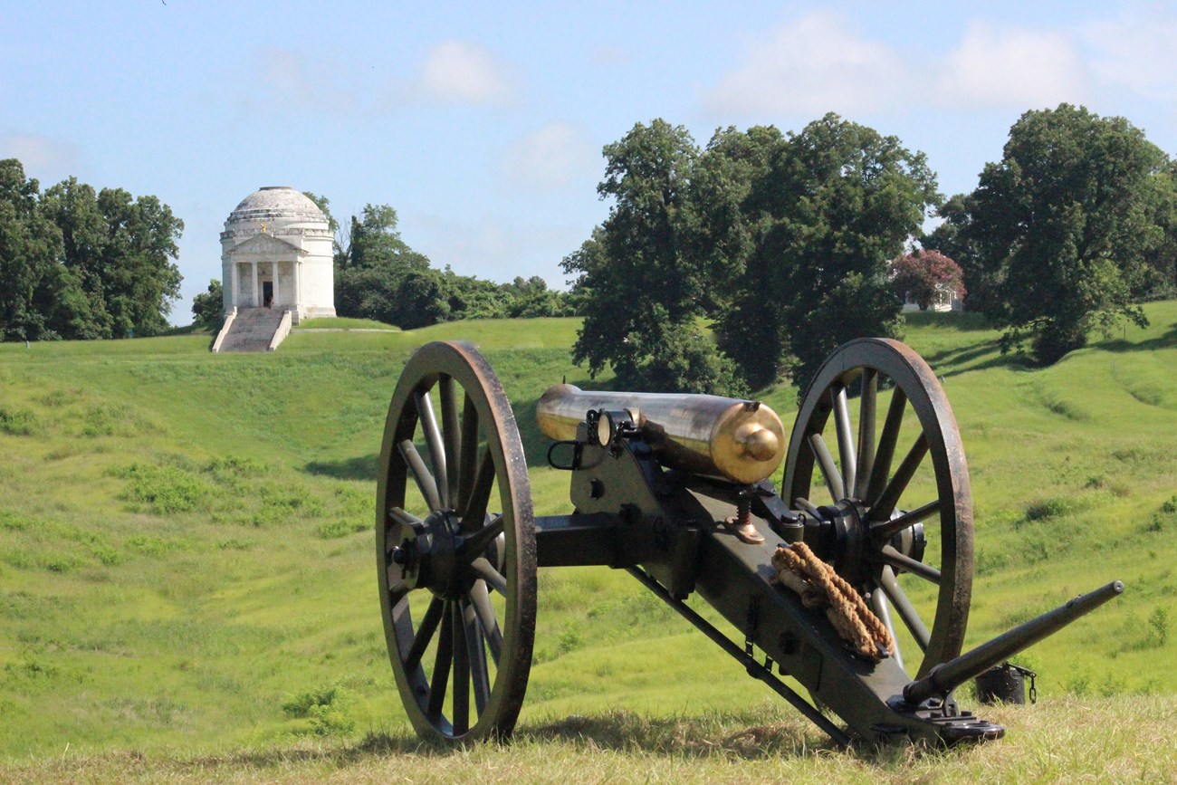 A bronze cannon facing a green field of grass and large white monument.